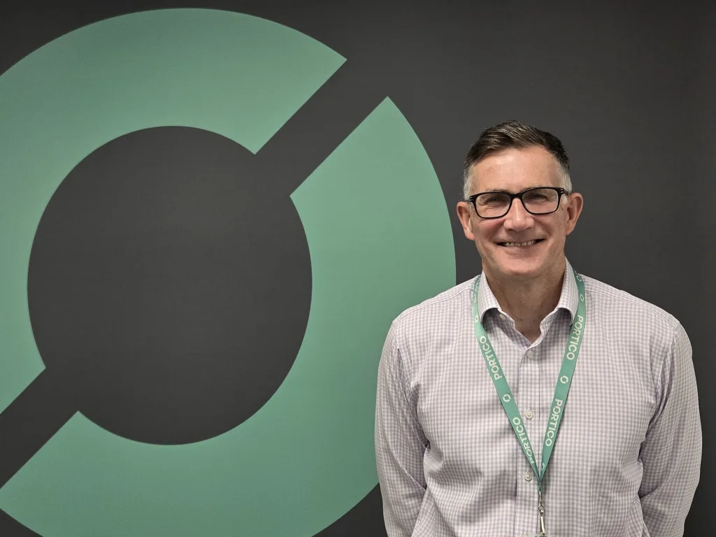 A picture of Simon Clarke, standing in front of a wall bearing the Portico shipping logo